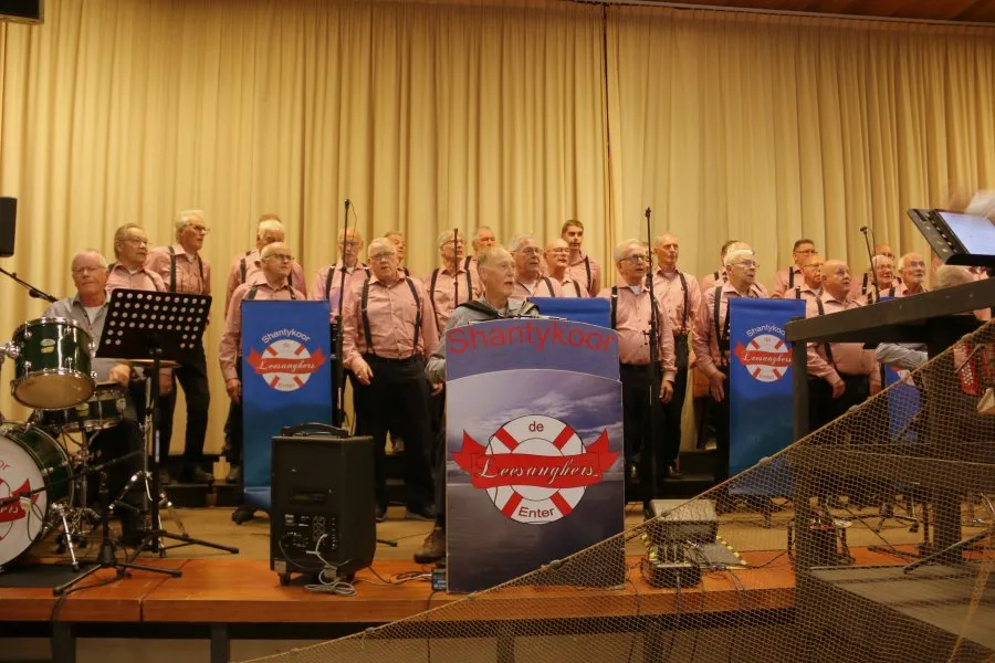 De Leesanghers op het podium, met drumstel, muziekstandaards en de koorborden met het reddingsboeilogo